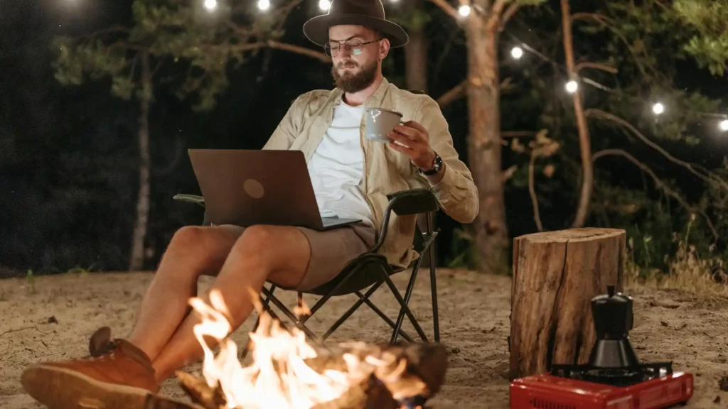 Um jovem com chapéu na frente de uma fogueira enquanto trabalha tranquilamente com seu notebook.