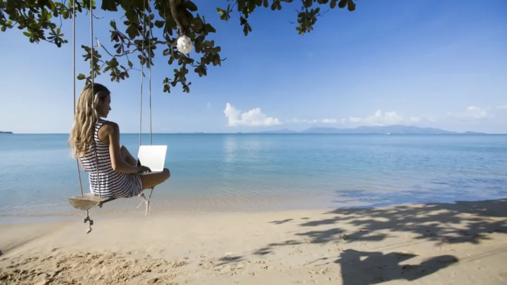 Um garota em uma rede na praia enquanto trabalha tranquilamente com seu notebook.